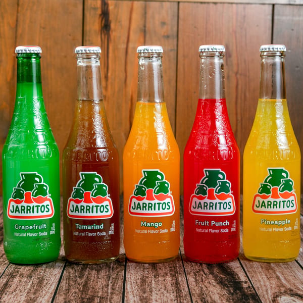 Five colorful Jarritos sodas in glass bottles, including grapefruit, tamarind, mango, fruit punch, and pineapple.
