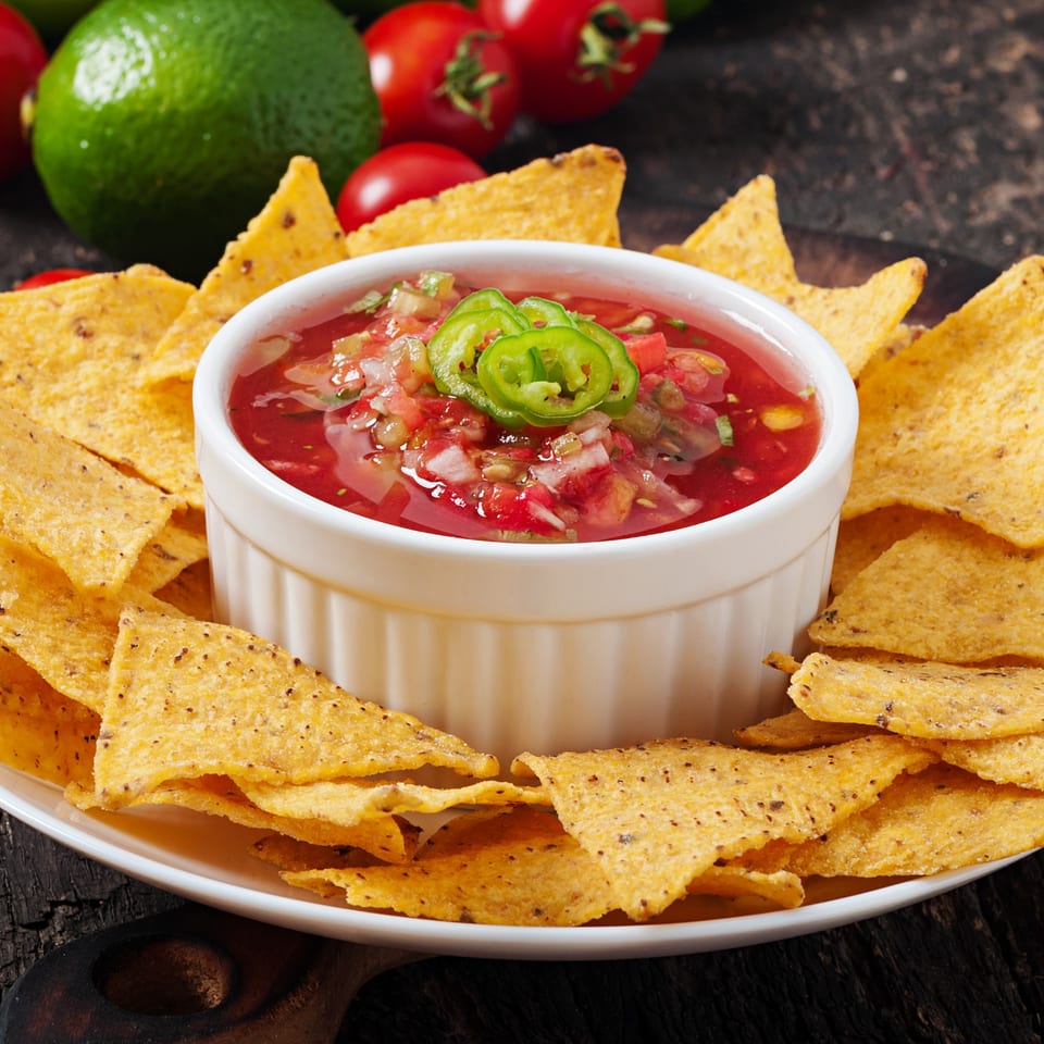 A white bowl of La Mesa salsa with jalapeños, surrounded by tortilla chips, limes, and tomatoes.