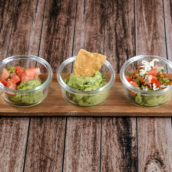 Three small glass bowls of guacamole with different toppings on a wooden board.